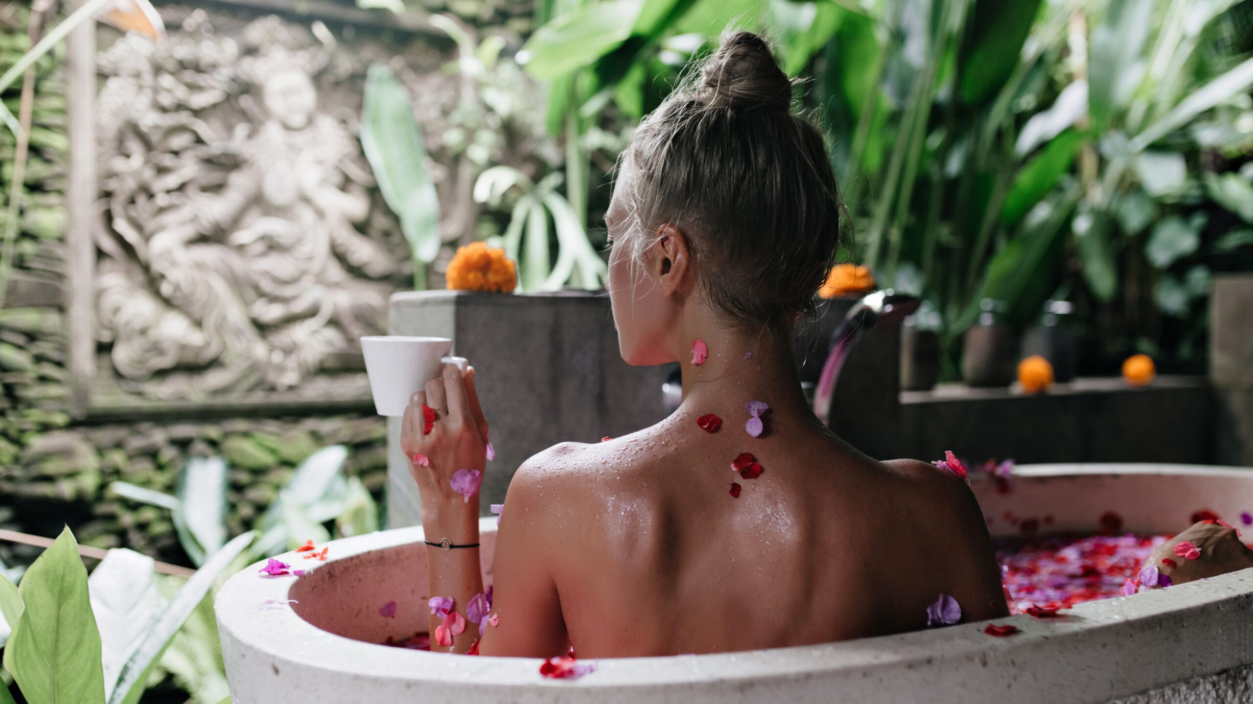 Woman relaxing in an outdoor stone bathtub with rose petals, enjoying a peaceful spa moment